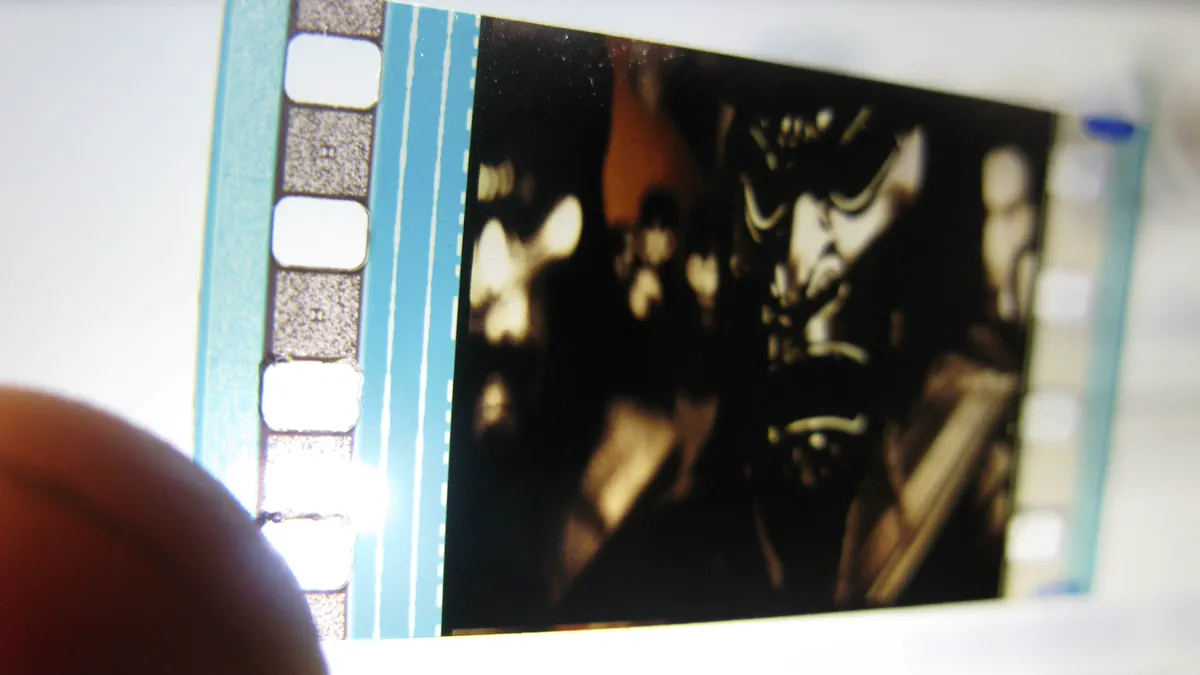 A strip of 35 mm film held up against a window, one dark frame lit from behind beside the pale sprocket holes, with a thumb out of focus in the lower corner.