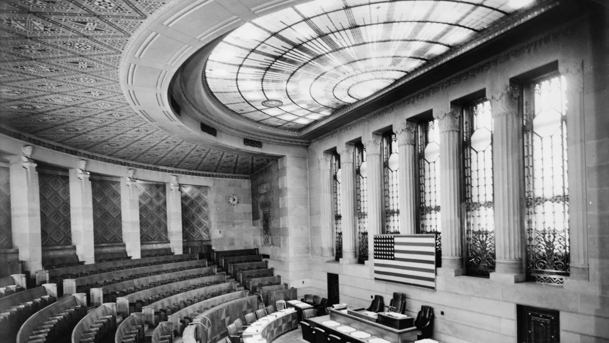 An empty council chamber in black and white: a curved desk with papers and microphones, tiered wooden benches, a flag on the wall, and a circular glass ceiling light above.