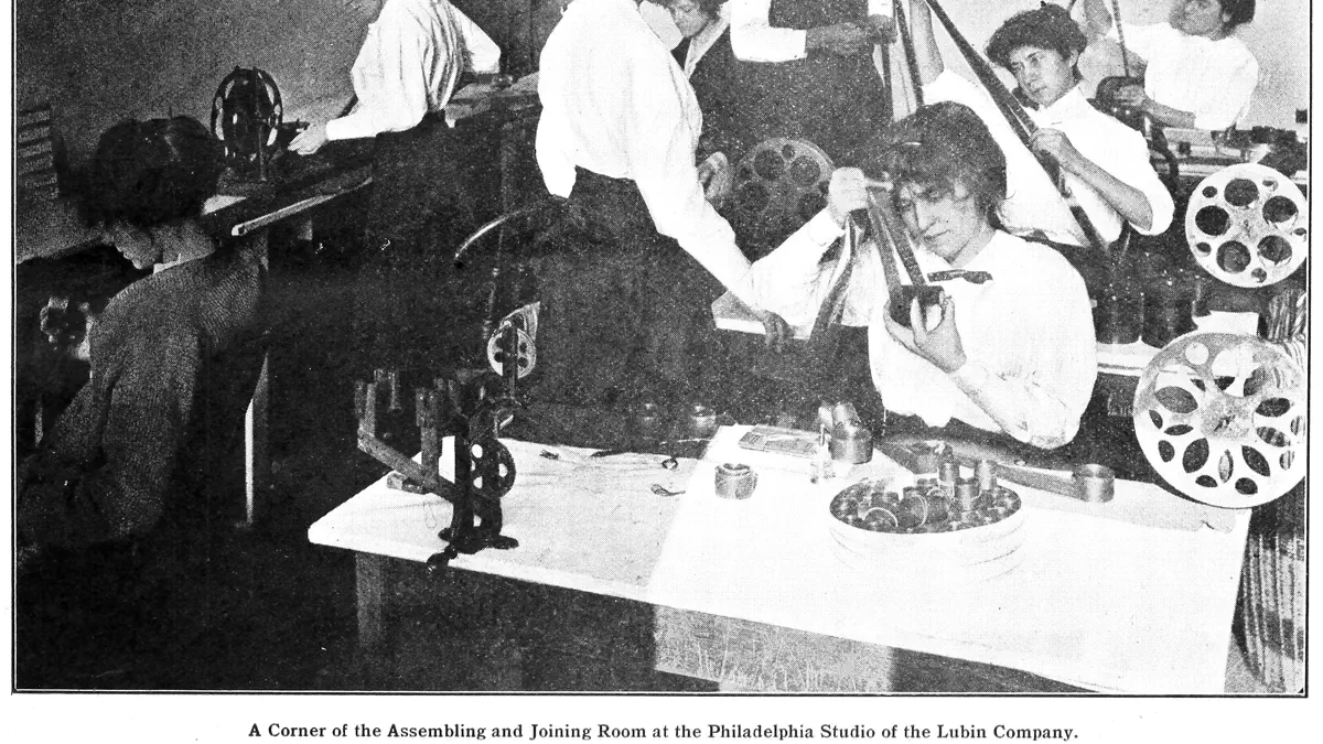 A black and white photograph of a film joining room: six women and a man at benches with reels of film, a splicer, strips of film hanging over the edges and small tins spread on a table.