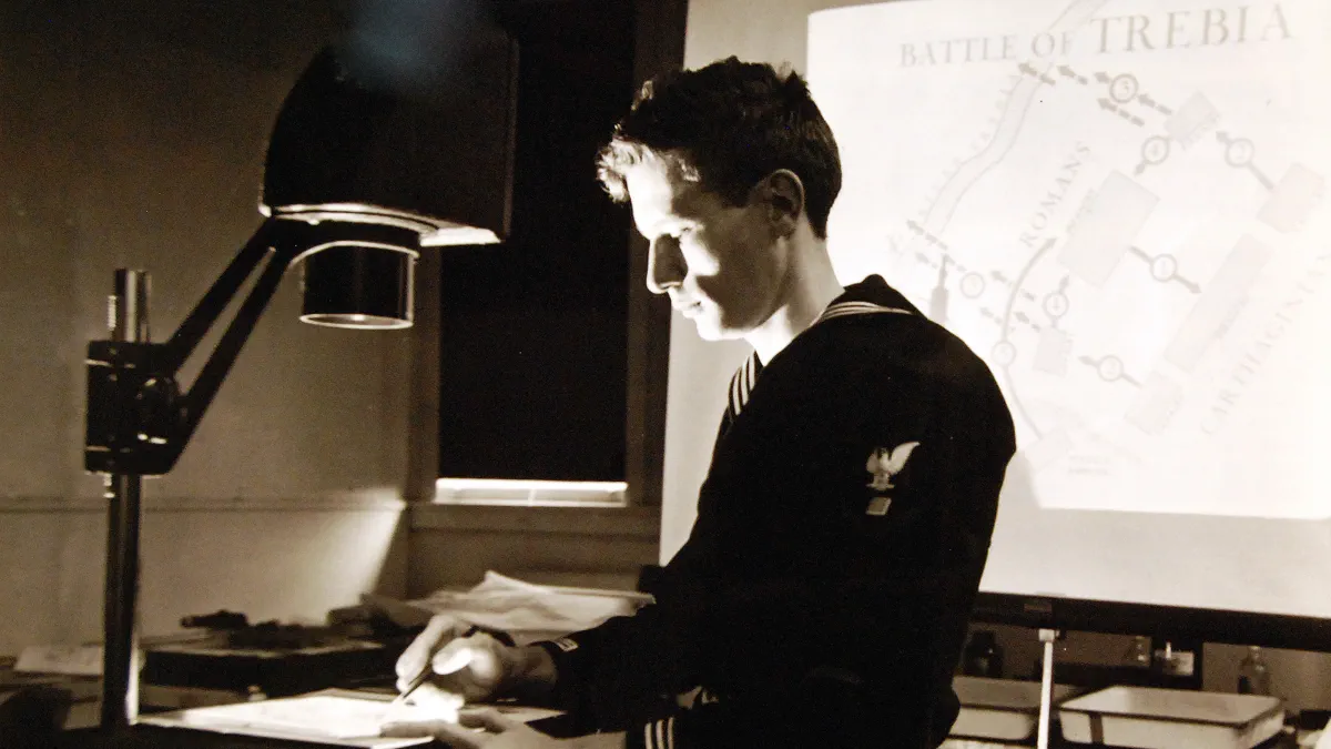 A sailor seated at a desk draws on a transparent sheet under the arm of a projector, while a map headed Battle of Trebia is thrown onto a screen behind him.