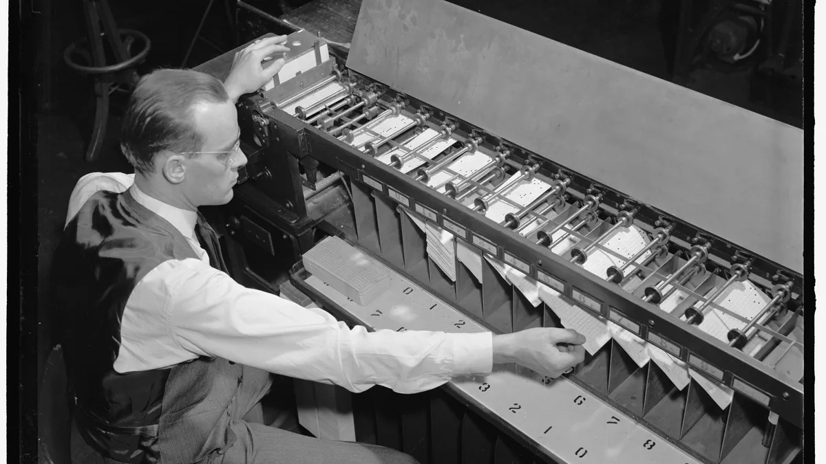 A man in a waistcoat feeds punched cards into a long mechanical sorter, which drops each card into one of twelve numbered pockets.