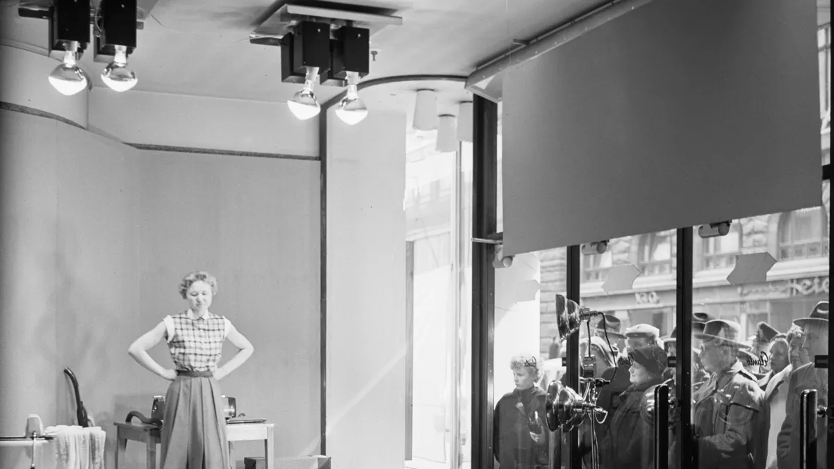 A woman in a checked blouse stands in a shop window beside laundry equipment while cameras on tripods film her and a crowd watches from the street through the glass.