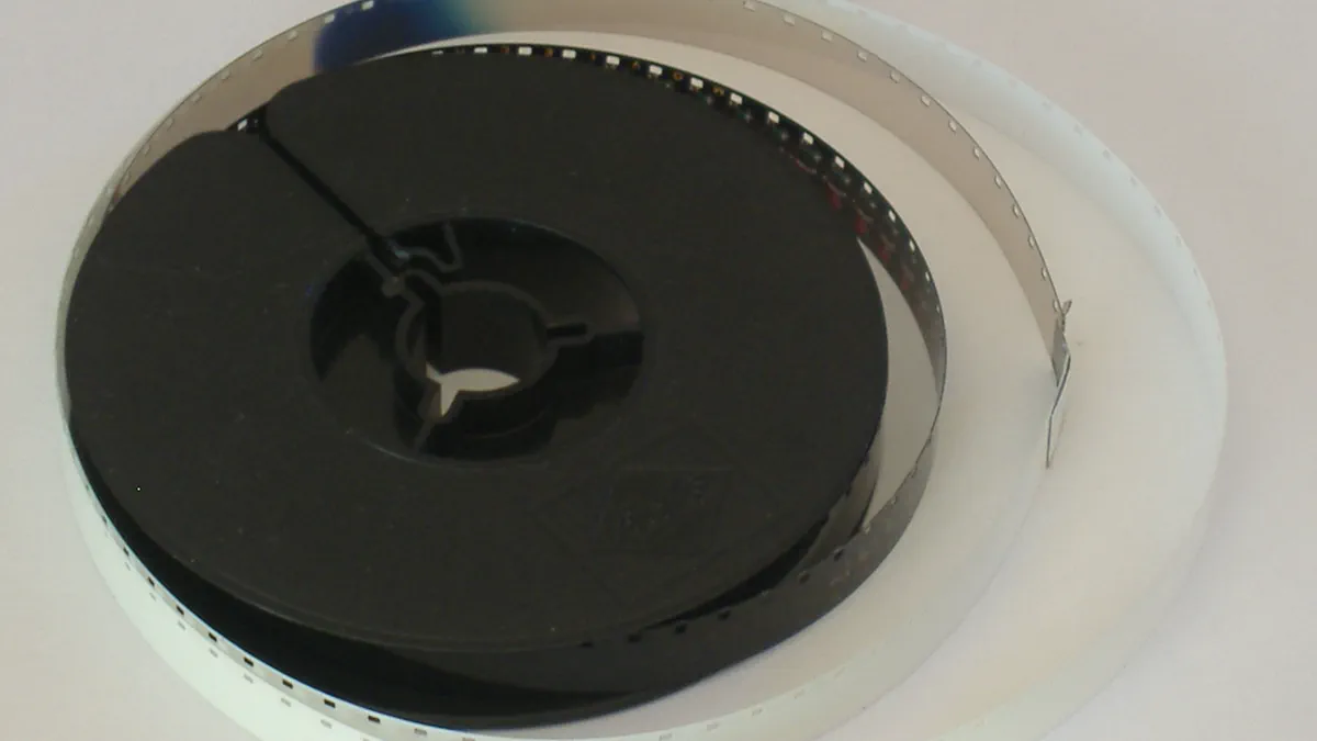 A Super 8 film reel lying on its side with part of the film unwound beside it, the sprocket holes visible along the edge of the black strip.