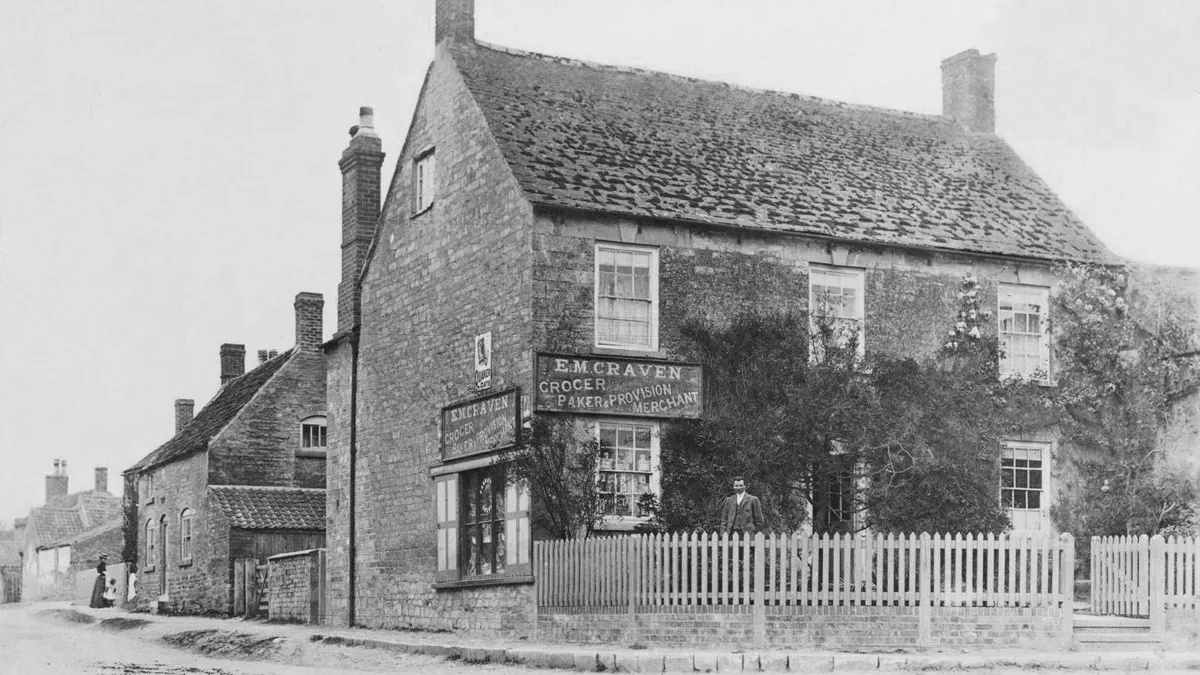 A stone village shop on a corner with two painted boards reading E. M. Craven, grocer, paker and provision merchant, a man standing at the gate and a woman walking past in the road.